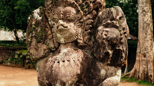Weathered stone sculpture of a seated figure surrounded by greenery, standing beside a pathway within a historic heritage site