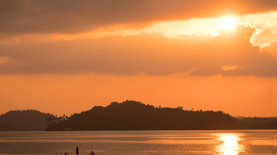 A vibrant sunset over a body of water, with a small boat silhouetted against the orange and yellow sky.