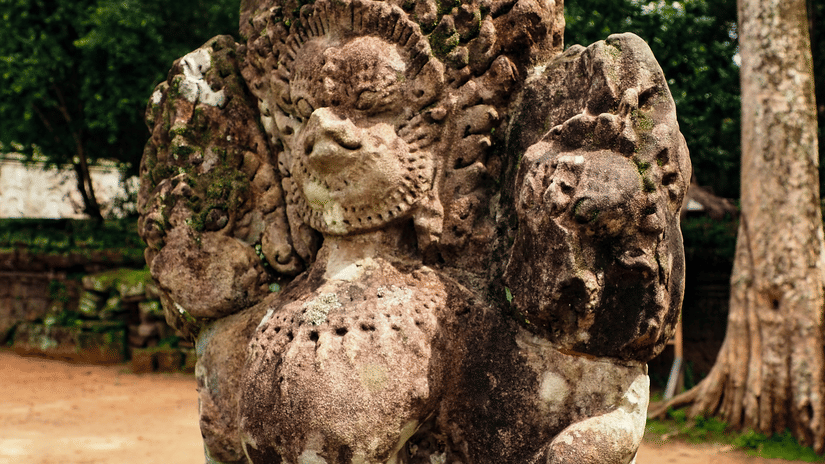 Weathered stone sculpture of a seated figure surrounded by greenery, standing beside a pathway within a historic heritage site