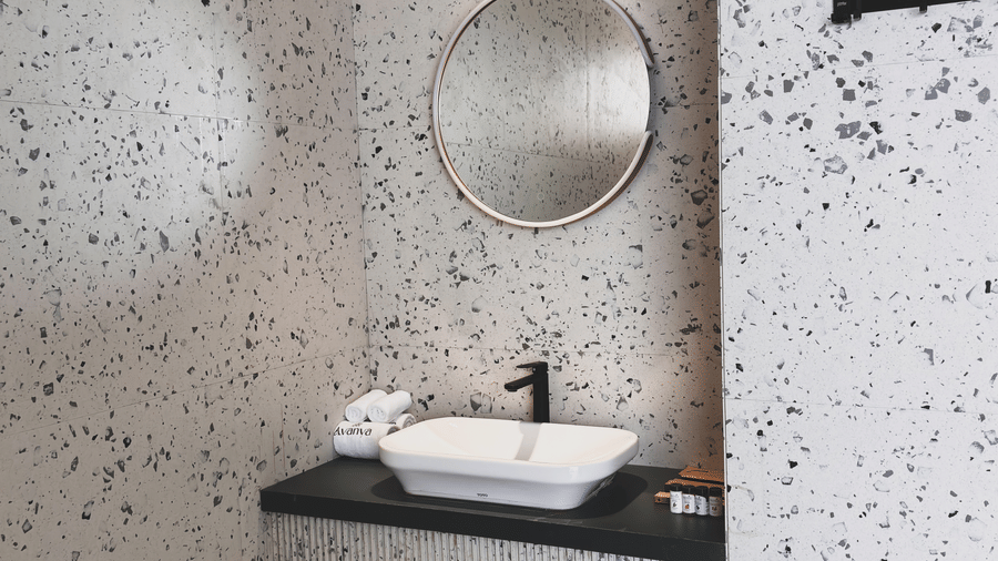 A white rectangular washbasin sits on a dark countertop against a grey-and-black speckled wall with a round mirror at Avanya Resorts N Retreat.