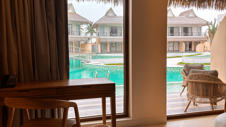 A wooden desk and chair stand by a large glass window offering a view of a pool and outdoor buildings at Avanya Resorts N Retreat.