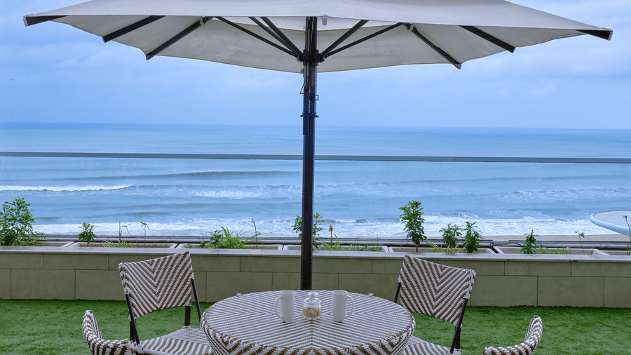 An outdoor patio of Premium Room at Swosti Premium Beach Resorts, Puri, set with a round table, four chairs, and a large, tiered umbrella on a grassy area overlooking the ocean under a cloudy sky.