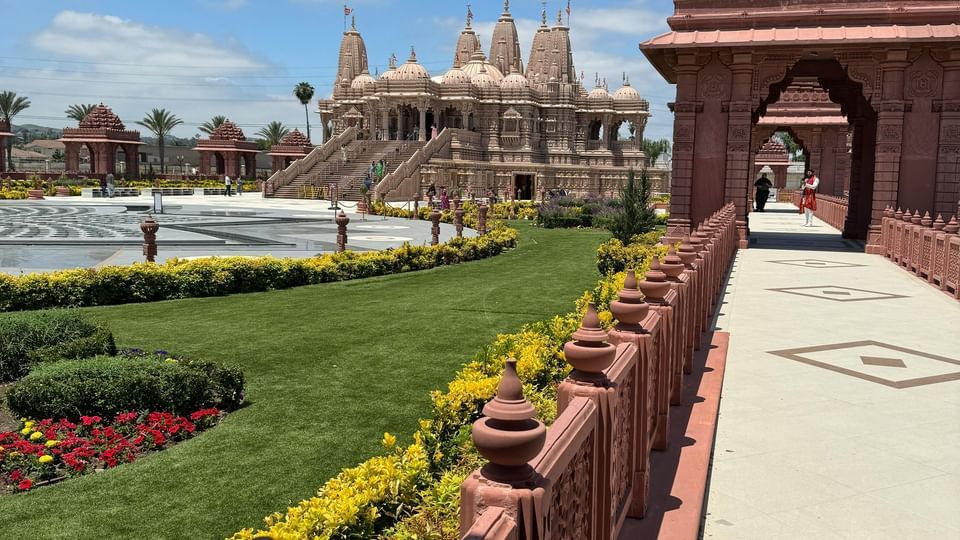 A wide, manicured walkway leads to a large, ornate temple entrance built from red stone under a bright blue, sunny sky.