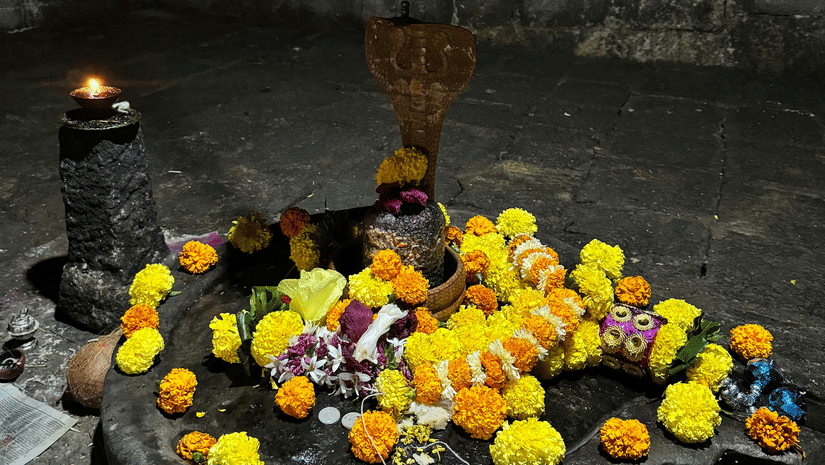 A Shiva lingam placed on a stone base with flowers arranged around it and a metal pot hanging above inside a temple area.