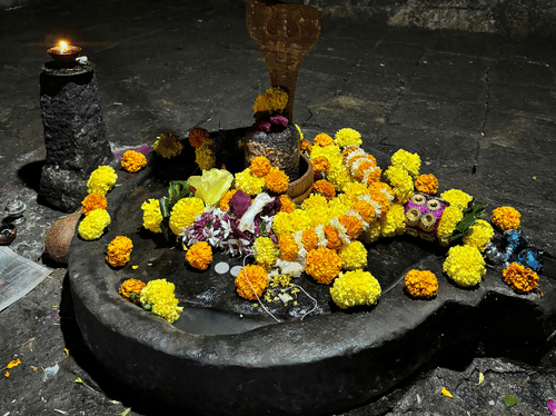 A Shiva lingam placed on a stone base with flowers arranged around it and a metal pot hanging above inside a temple area.