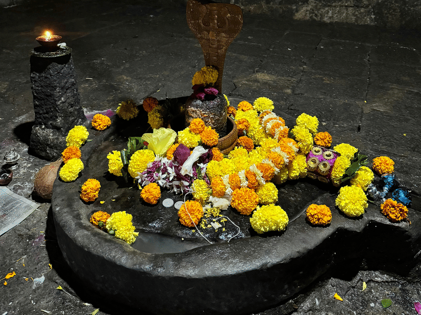A Shiva lingam placed on a stone base with flowers arranged around it and a metal pot hanging above inside a temple area.