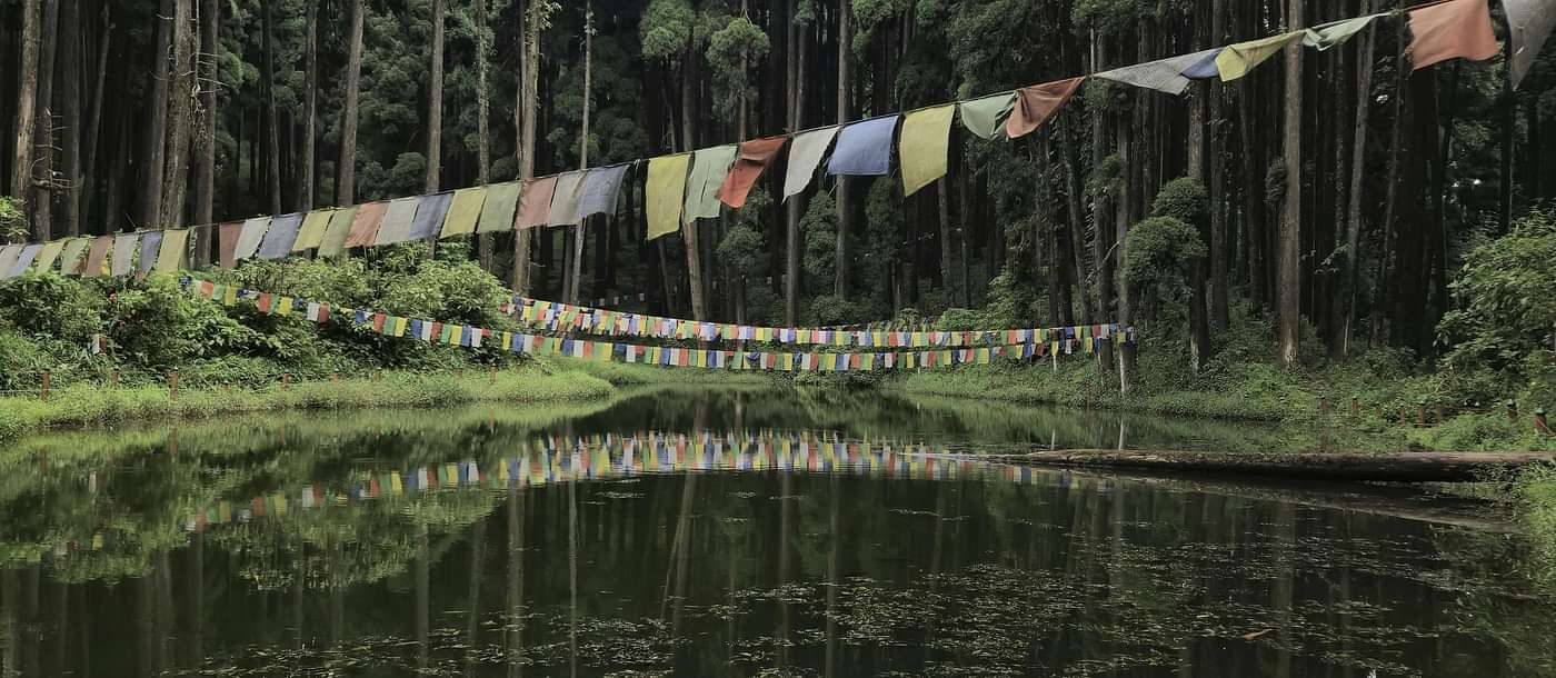 A still pond reflects tall trees and colourful prayer flags strung across its banks.
