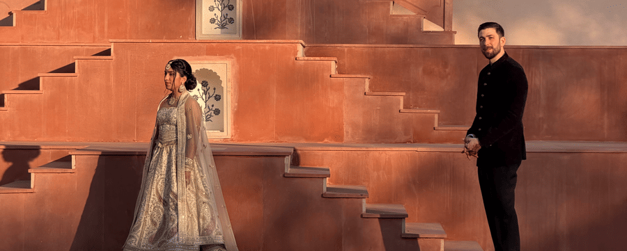 A bride stands on the wide stone steps while the groom looks up at her from below against the traditional red-stone architecture of Umaid Palace.