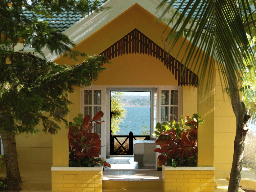 A building entrance with a triangular roof, plants near the doorway, and palm trees around the pathway leading to an open seating area inside.