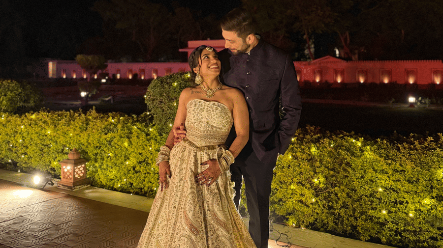 A bride and groom posing together on an outdoor lawn at night with the illuminated structure of Umaid Palace visible in the background.