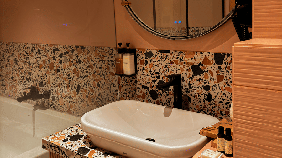 A large round mirror hangs above a white sink on a speckled countertop next to a white bathtub at Avanya Resorts N Retreat.