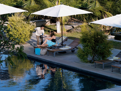 A woman lounges by a pool while a server offers her a drink. White umbrellas and tall palm trees surround the pool, with a lush green forest in the background, bathed in gentle sunlight - ??The Serai Chikmagalur