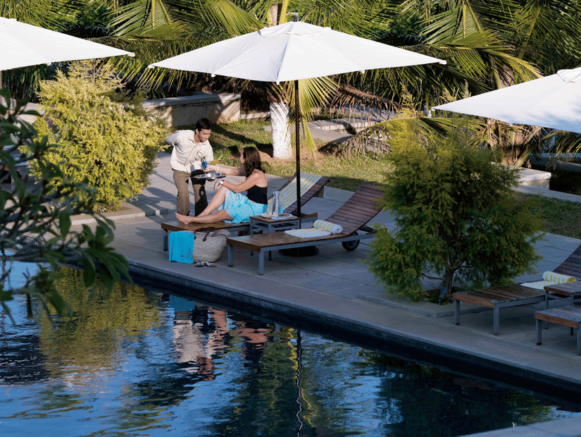 A woman lounges by a pool while a server offers her a drink. White umbrellas and tall palm trees surround the pool, with a lush green forest in the background, bathed in gentle sunlight - ??The Serai Chikmagalur