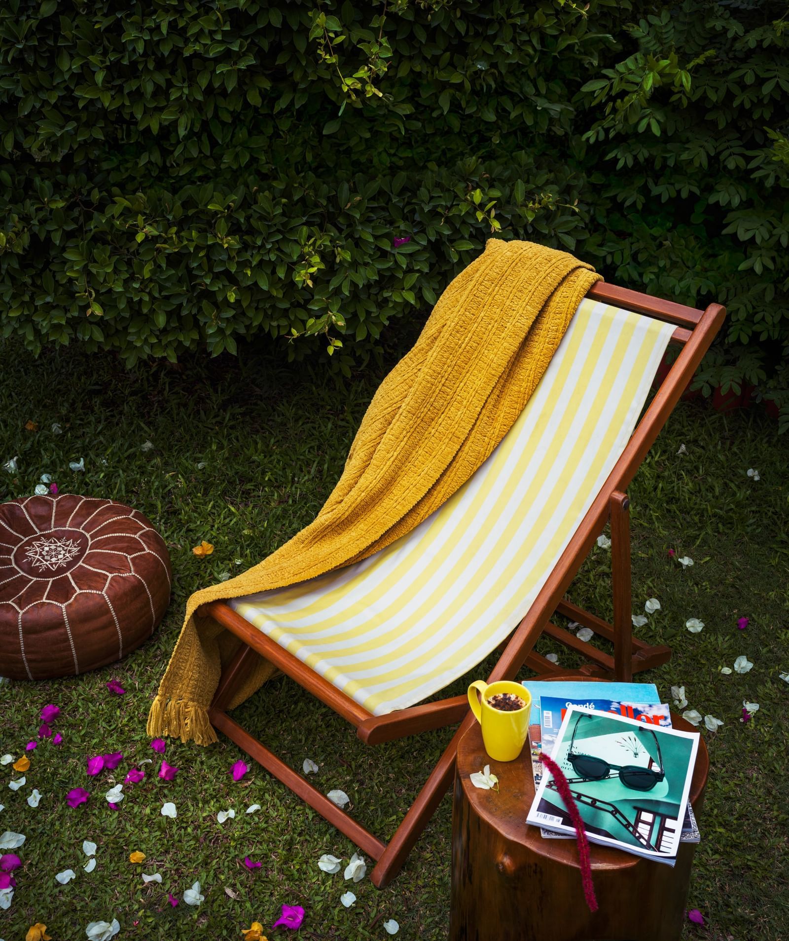 A yellow and white striped deck chair with a mustard towel placed over it on a green lawn with a small side table besides it.