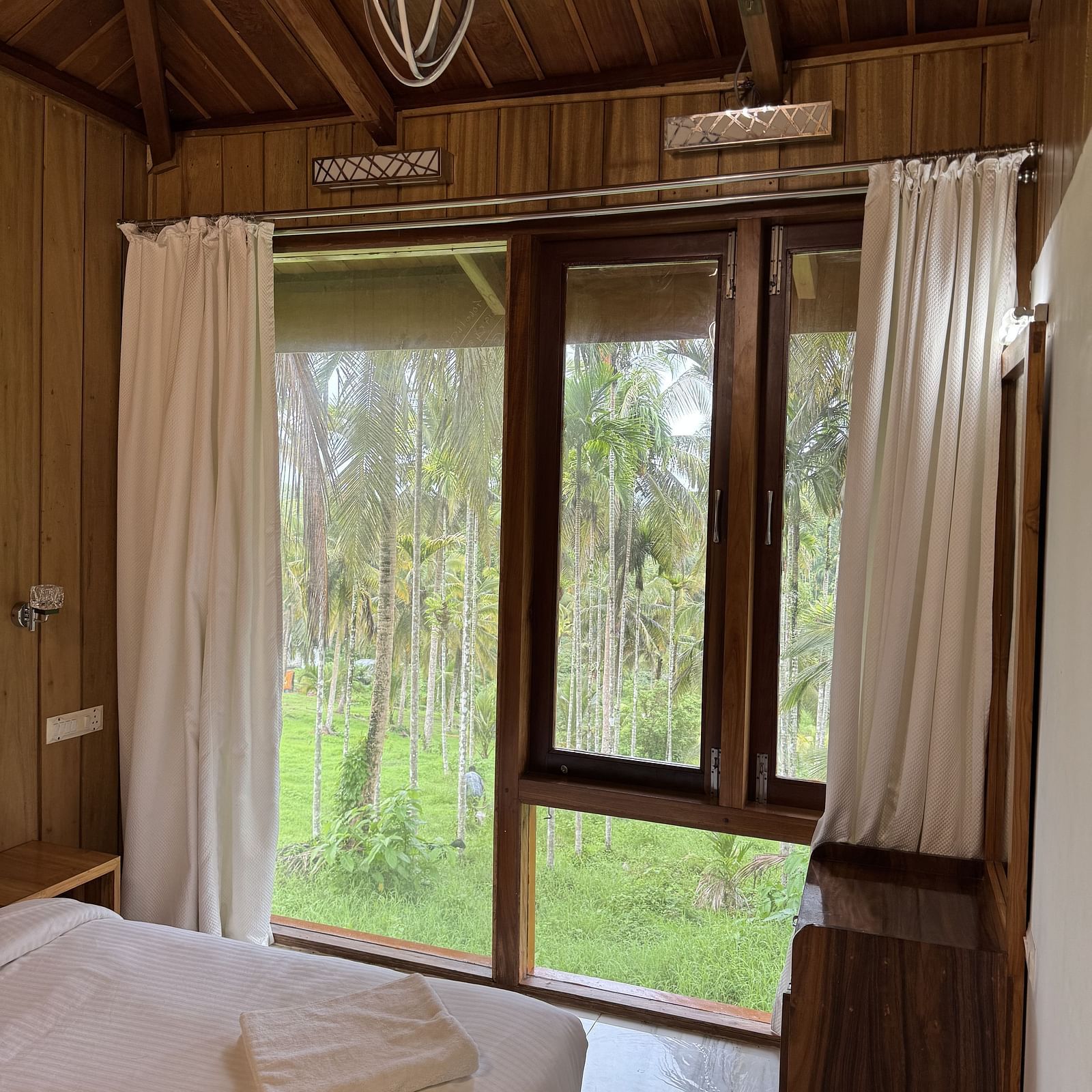 A resort bedroom with two beds, wooden walls and ceiling, a ceiling fan, and large windows with curtains.