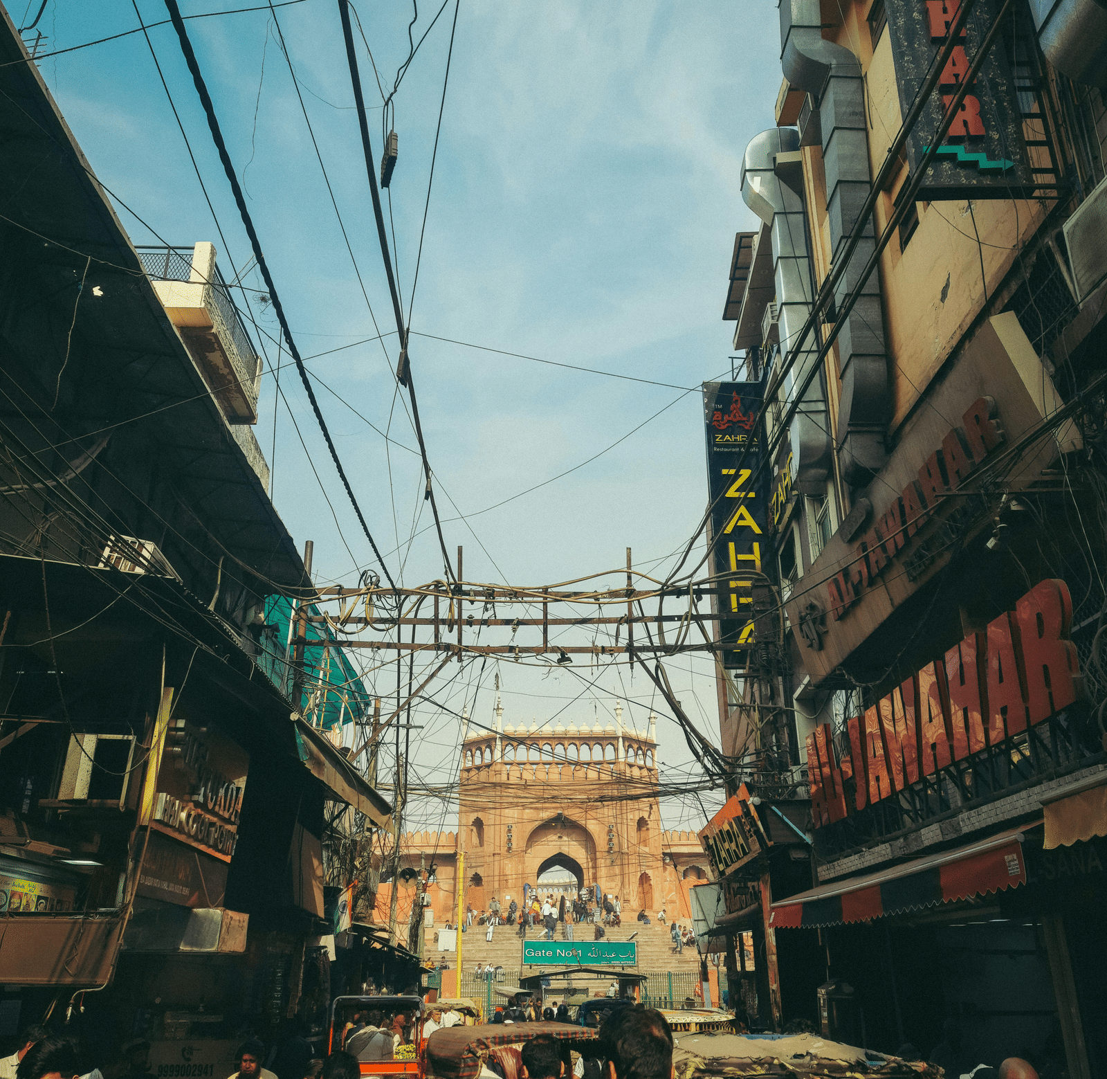 Busy street market scene in India with people, buildings, and overhead wires.
