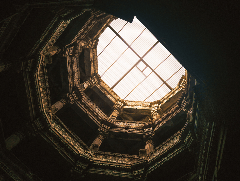 A view looking up from the dark depths of an ancient octagonal stepwell towards a bright, light-filled opening.