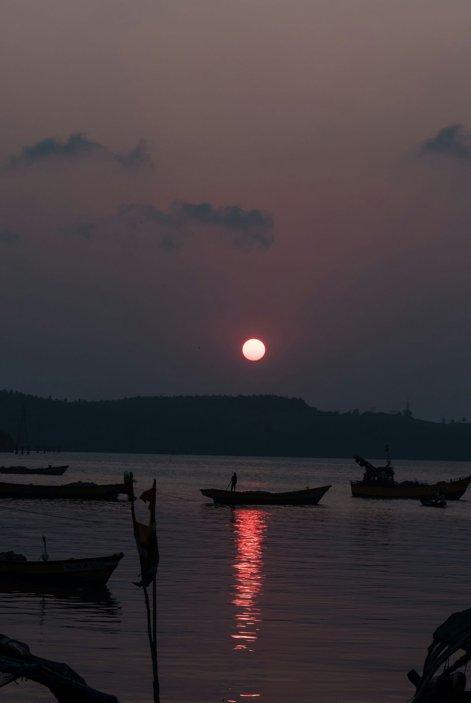 A tranquil sunset with a deep red sun reflecting on the water, silhouetting fishing boats in the foreground