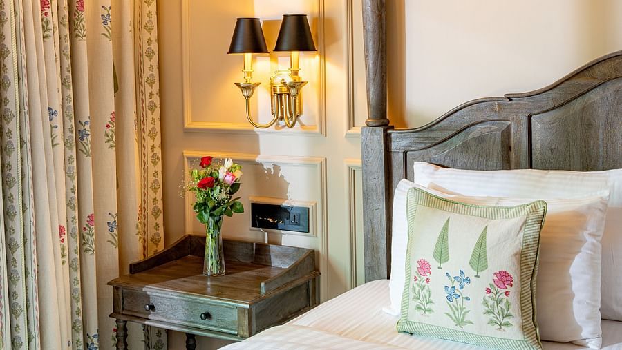 Cosy corner of a hotel bedroom featuring a wooden four-poster bed with floral cushions, a vintage wooden nightstand with a vase of fresh flowers, elegant wall sconces, floral curtains, and a classic chandelier above - Panna Vilas Palace