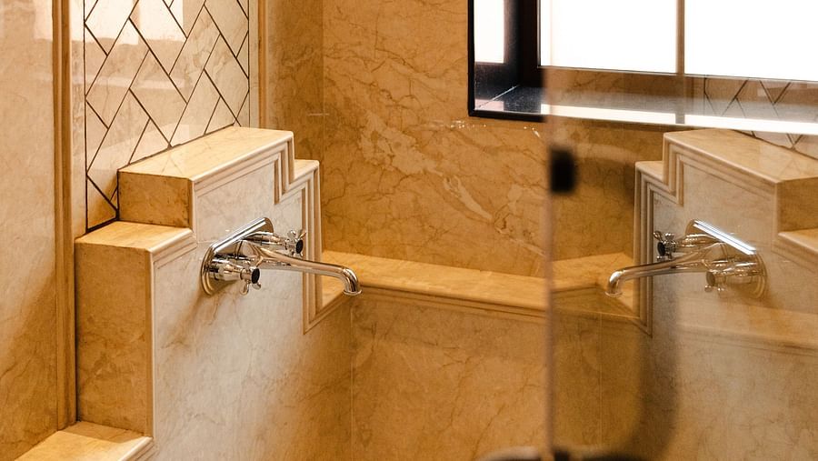 Elegant bathroom interior featuring beige marble walls, a glass partition, a window with frosted panes, and twin chrome wall-mounted faucets above a built-in stone ledge - Panna Vilas Palace