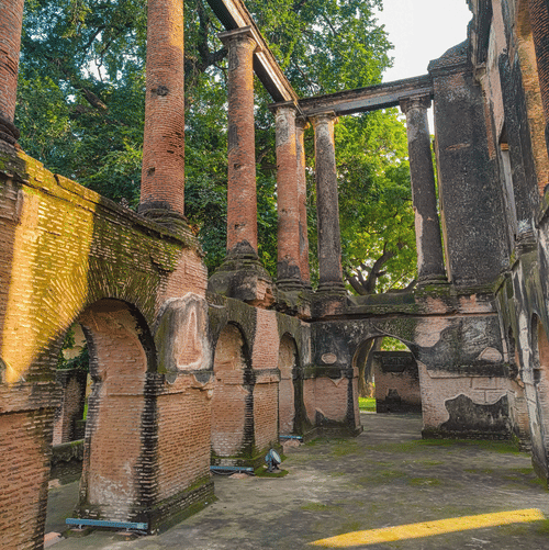 The ruins of The Residency, Lucknow. There are tall walls with arched doorways, the floor is covered in moss and trees are visible at the back.