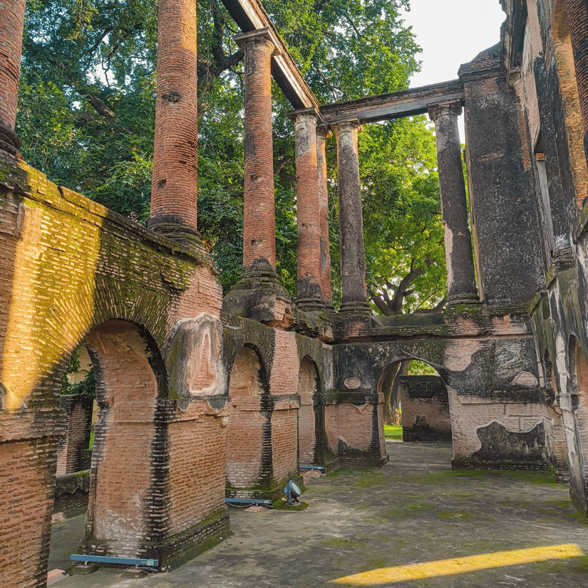 The ruins of The Residency, Lucknow. There are tall walls with arched doorways, the floor is covered in moss and trees are visible at the back.