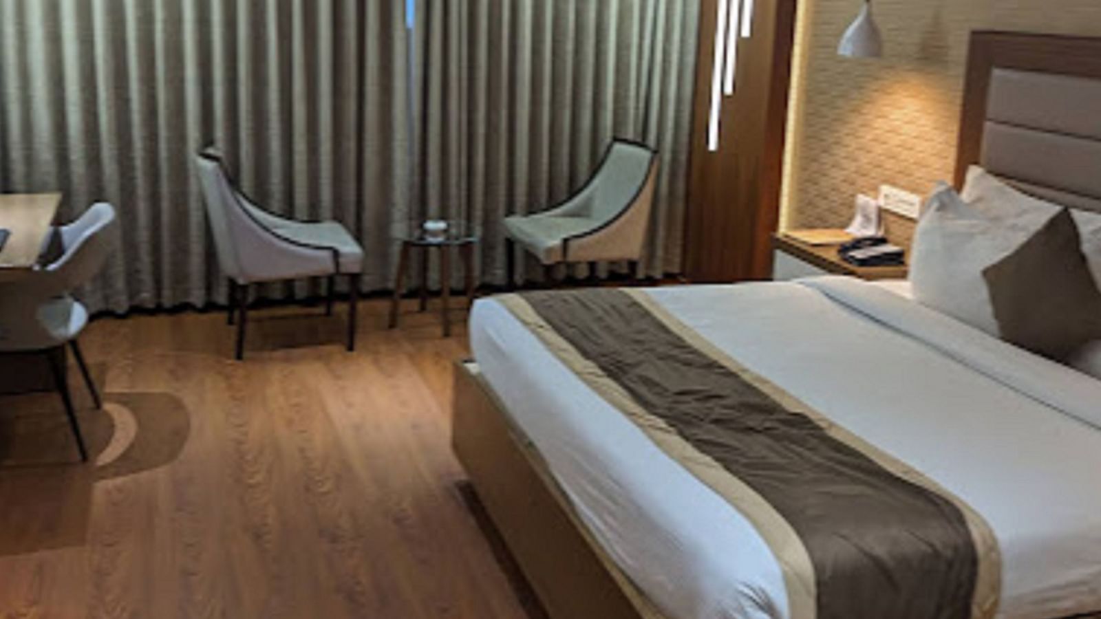 A premium hotel room with a neatly made large bed, windows with closed curtains, along with two chairs at Yashshree The Imperial Inn, Siliguri.