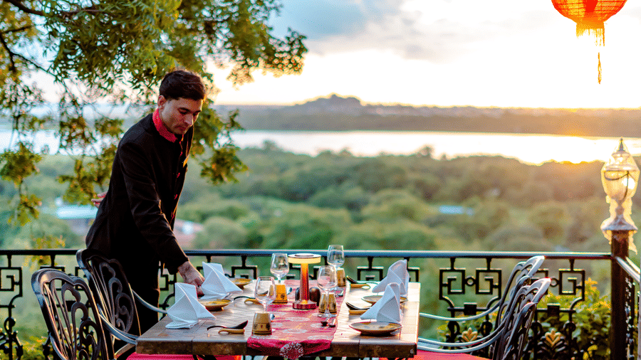 An outdoor dining table set for a meal at Ming Dynasty, part of the Noor-Us-Sabah Palace, Bhopal, overlooking a scenic lake view at sunset with a red lantern hanging nearby.