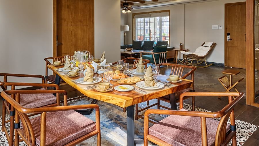 Stylish dining area with a wooden table set for six, surrounded by mid-century chairs on patterned tiles. Gold pendant lights hang from a recessed ceiling, with a cosy living space visible in the background - Voyage The Solitaire Bliss.