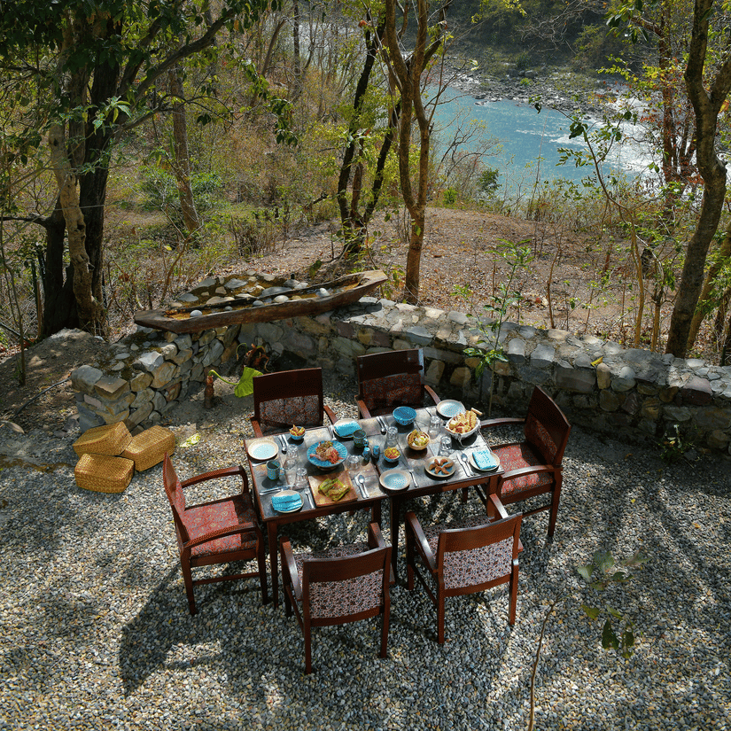 Scenic outdoor dining area with a wooden table and chairs set amongst tall trees with a glimpse of the river in the background at Neemrana’s Glasshouse on The Ganges creating a unique and tranquil setting for alfresco meals in the heart of nature