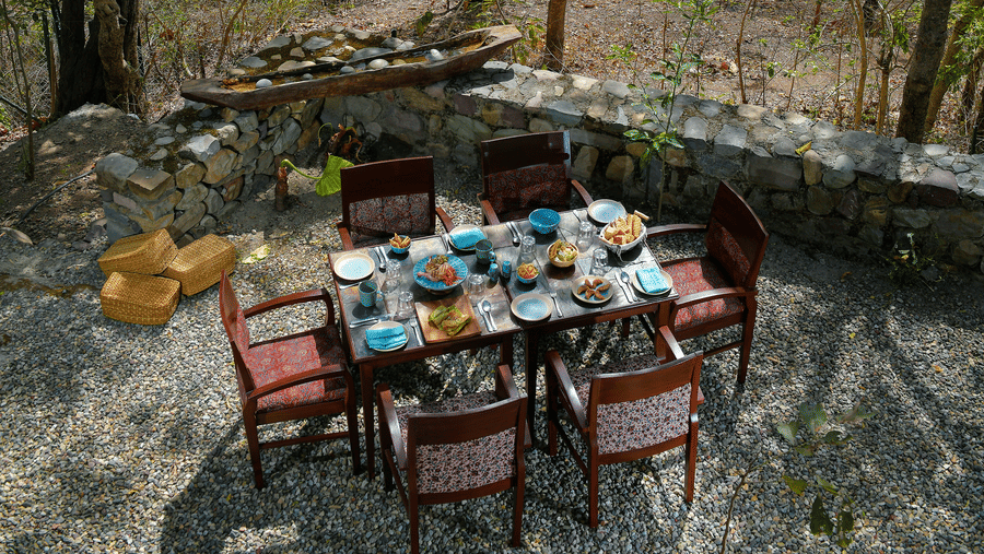 Scenic outdoor dining area with a wooden table and chairs set amongst tall trees with a glimpse of the river in the background at Neemrana’s Glasshouse on The Ganges creating a unique and tranquil setting for alfresco meals in the heart of nature