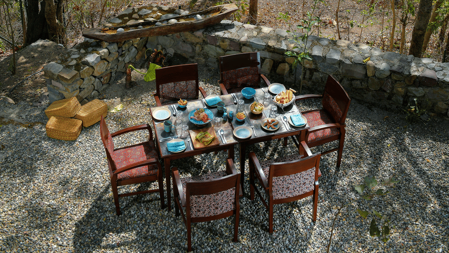Scenic outdoor dining area with a wooden table and chairs set amongst tall trees with a glimpse of the river in the background at Neemrana’s Glasshouse on The Ganges creating a unique and tranquil setting for alfresco meals in the heart of nature
