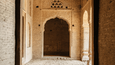 A view through a doorway into an old stone corridor featuring a carved arched entrance, textured brick walls, and arched openings along the side.