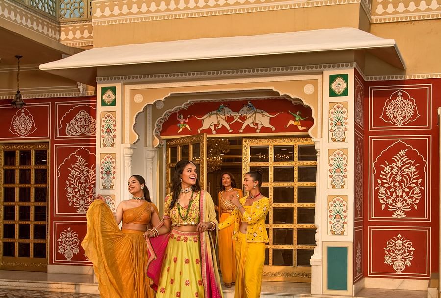 Three women in traditional indian attire stand in front of a colorful, ornate building with an arched entrance. they appear to be engaged in a cheerful conversation.
