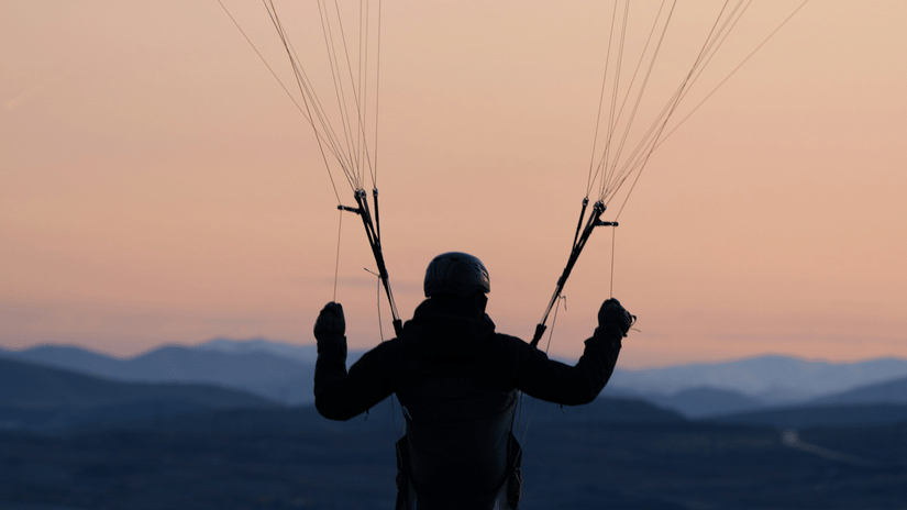 Paraglider silhouetted against a pastel sunset, descending over layered mountain ridges with arms raised during an evening Vagamon paragliding above mountains