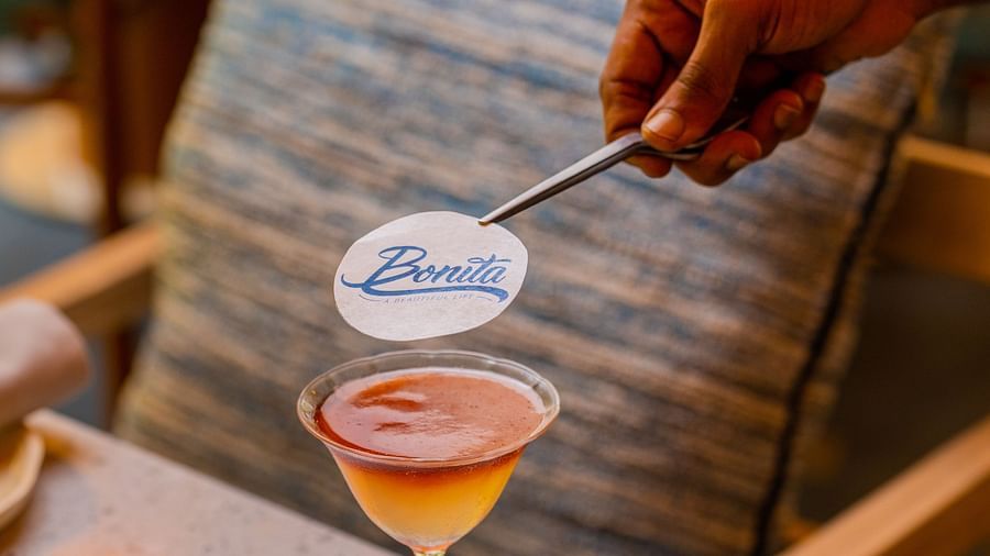 A close-up of a hand placing a Bonita-branded stirrer into a cocktail glass on a table, with a blurred background of cozy seating.