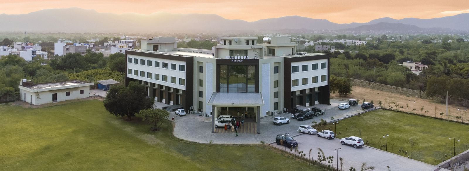 An aerial view of the Bamboo Saa Mulberry Resort, Pushkar at sunset or sunrise surrounded by green lawn and the backdrop of hills.