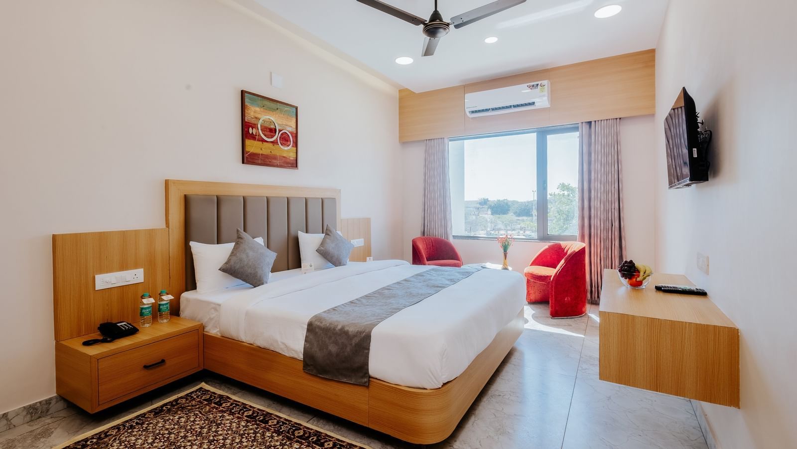 A spacious bedroom at Daksh The Grand Gowardhan, Dwarka, featuring a comfortable double bed opposite to a wall mounted television, and a common seating area at the far end of the room.