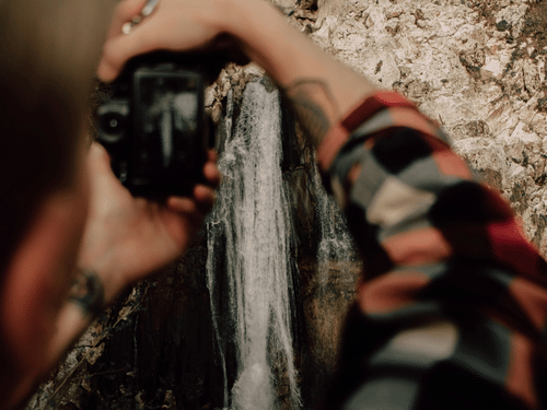 A person in a checked shirt photographing a small waterfall cascading over mossy rocks in a forest.