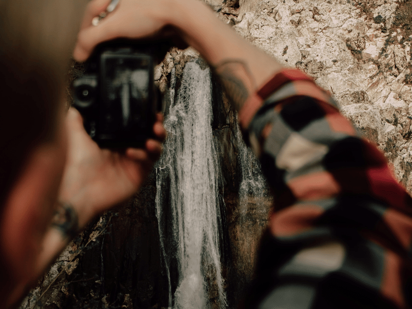A person in a checked shirt photographing a small waterfall cascading over mossy rocks in a forest.