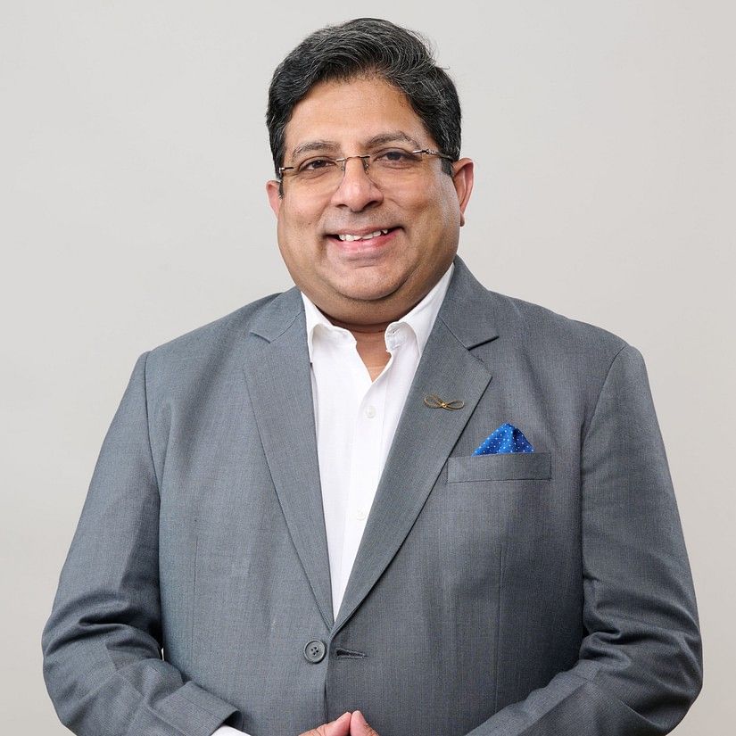 Debashish Chatterjee in a grey suit with a blue pocket square, standing with hands clasped, exuding confidence against a plain background.