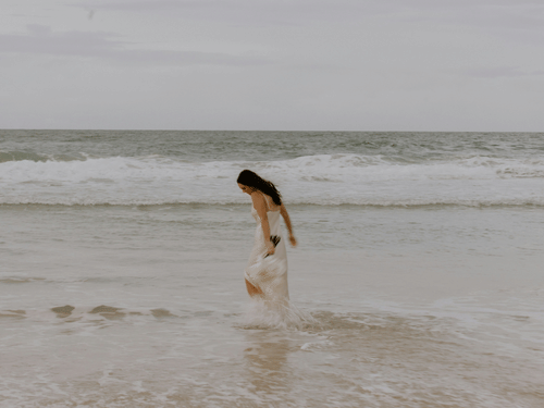 A woman in bridal dress walking barefoot along a quiet beach shoreline on an overcast day.