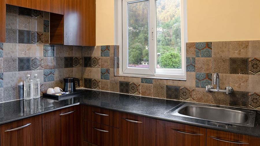 Kitchen features wooden cabinets, a dark worktop with a sink and tap, patterned splashback tiles, and a window showing a leafy outdoor view at Clarks Inn, Near Kainchi Dham.