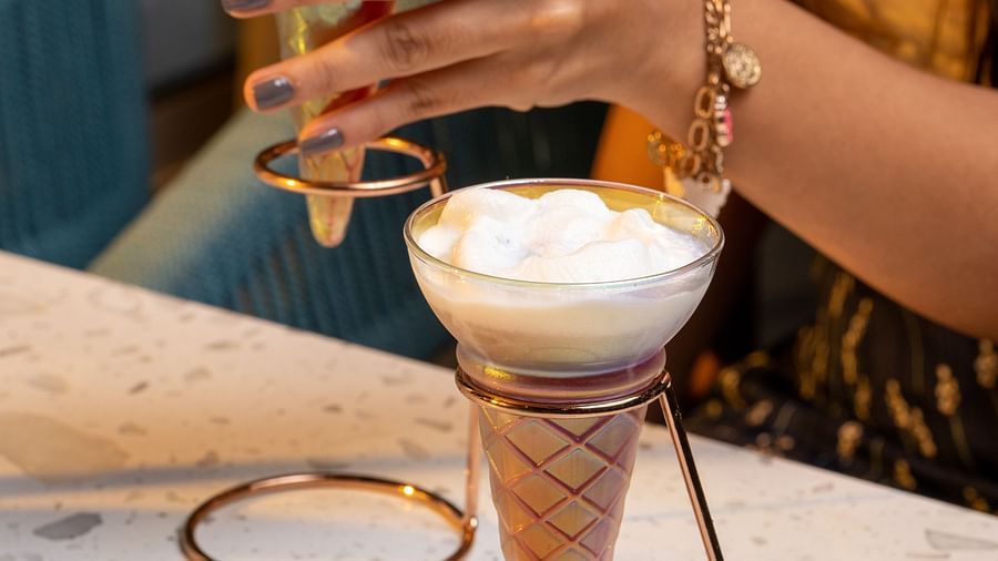 A close-up of an ice-cream cone shaped drink placed on a white table top and another one being prepared in the background.
