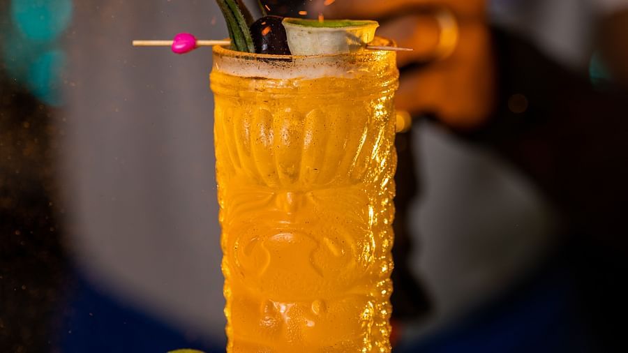 A close-up of a cocktail glass on a table which has orange coloured drink, with a blurred background of a flash lighter spray on the top of the glass..