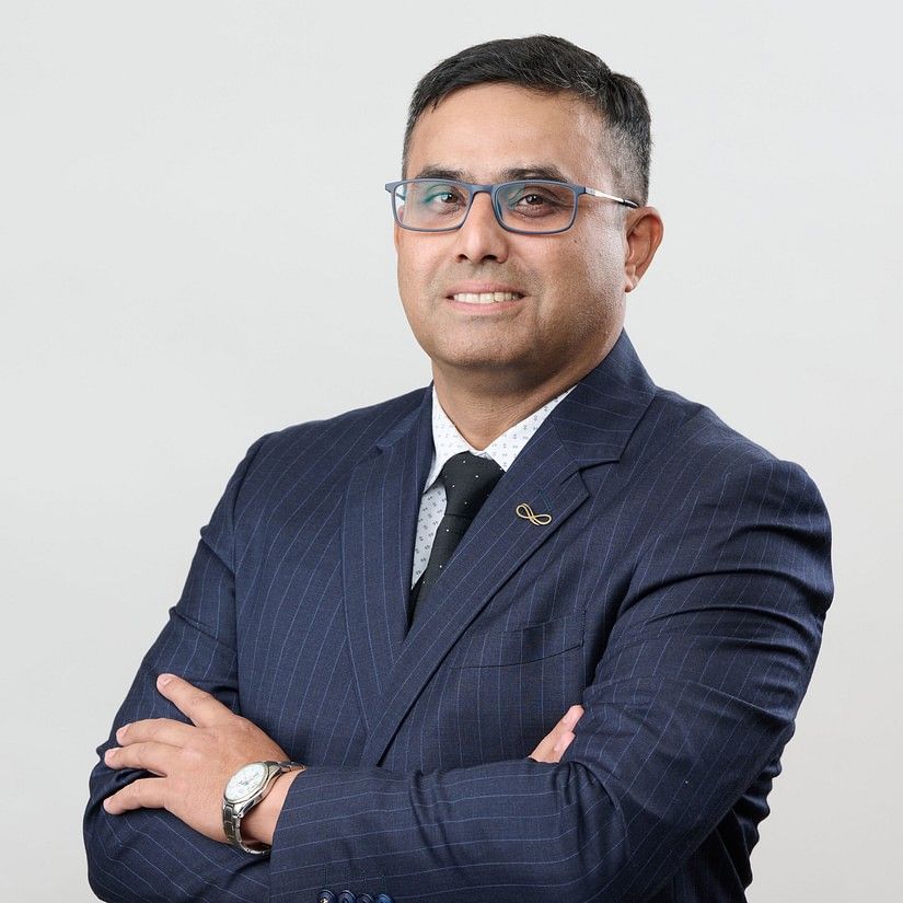 Siddhartha  in a DARK suit with a dark tie, wearing glasses stands with arms crossed, exuding confidence.