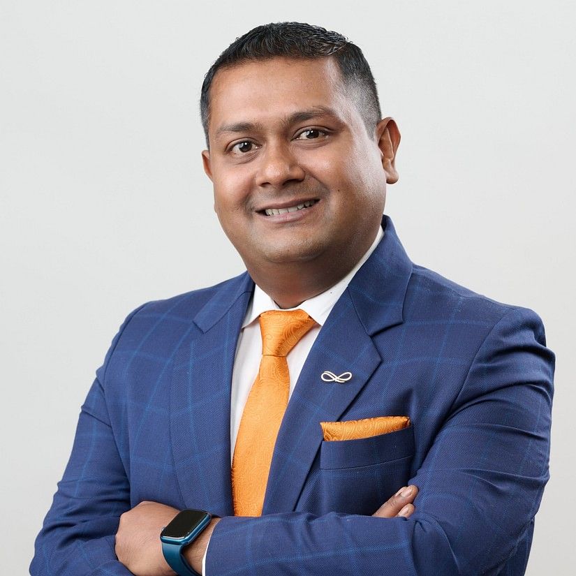 Suranjit Sengupta in a blue suit with a orange pocket square and tie stands with arms crossed, exuding confidence.