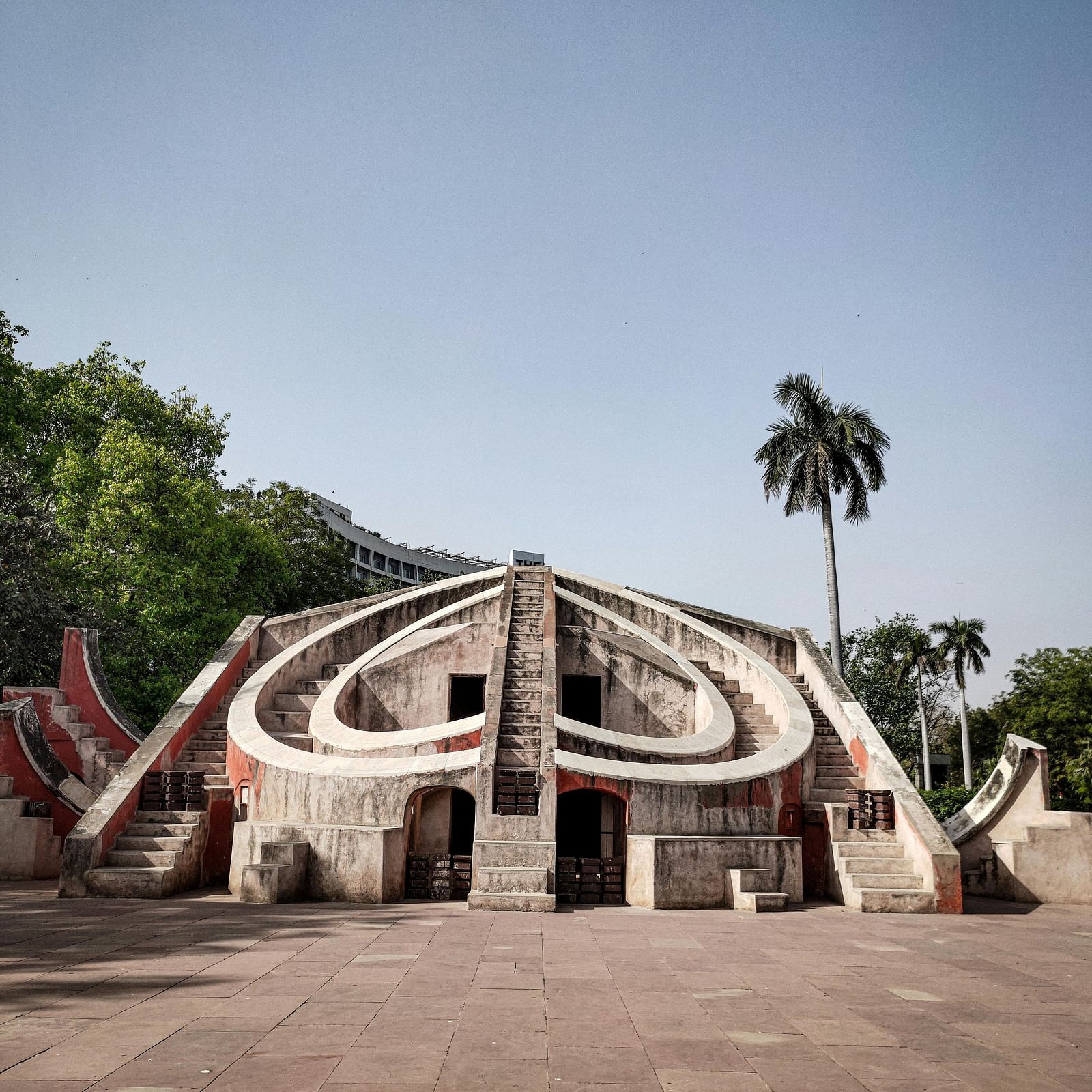 An astronomical instrument at Jantar Mantar, features multiple concrete structures with curved surfaces and openings under a clear sky, surrounded by trees and a palm tree.