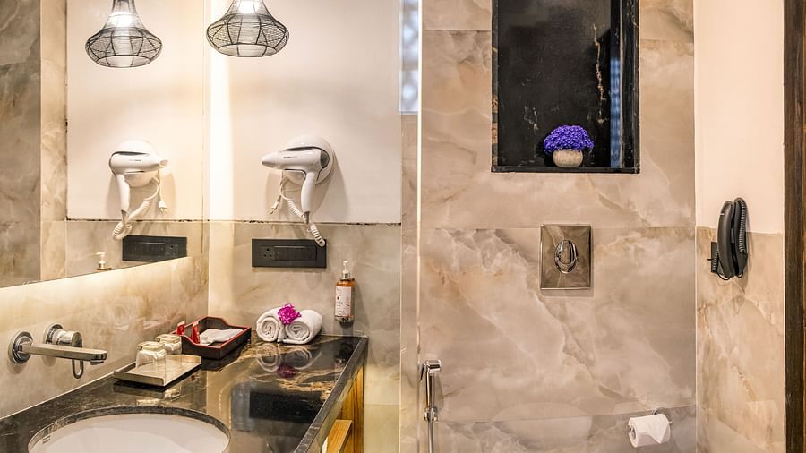 A modern bathroom with a dark countertop, a vessel sink, and hanging pendant lights. It features a white toilet, a small window, and marble-like tile walls - Lords Eco Inn Dehradun.