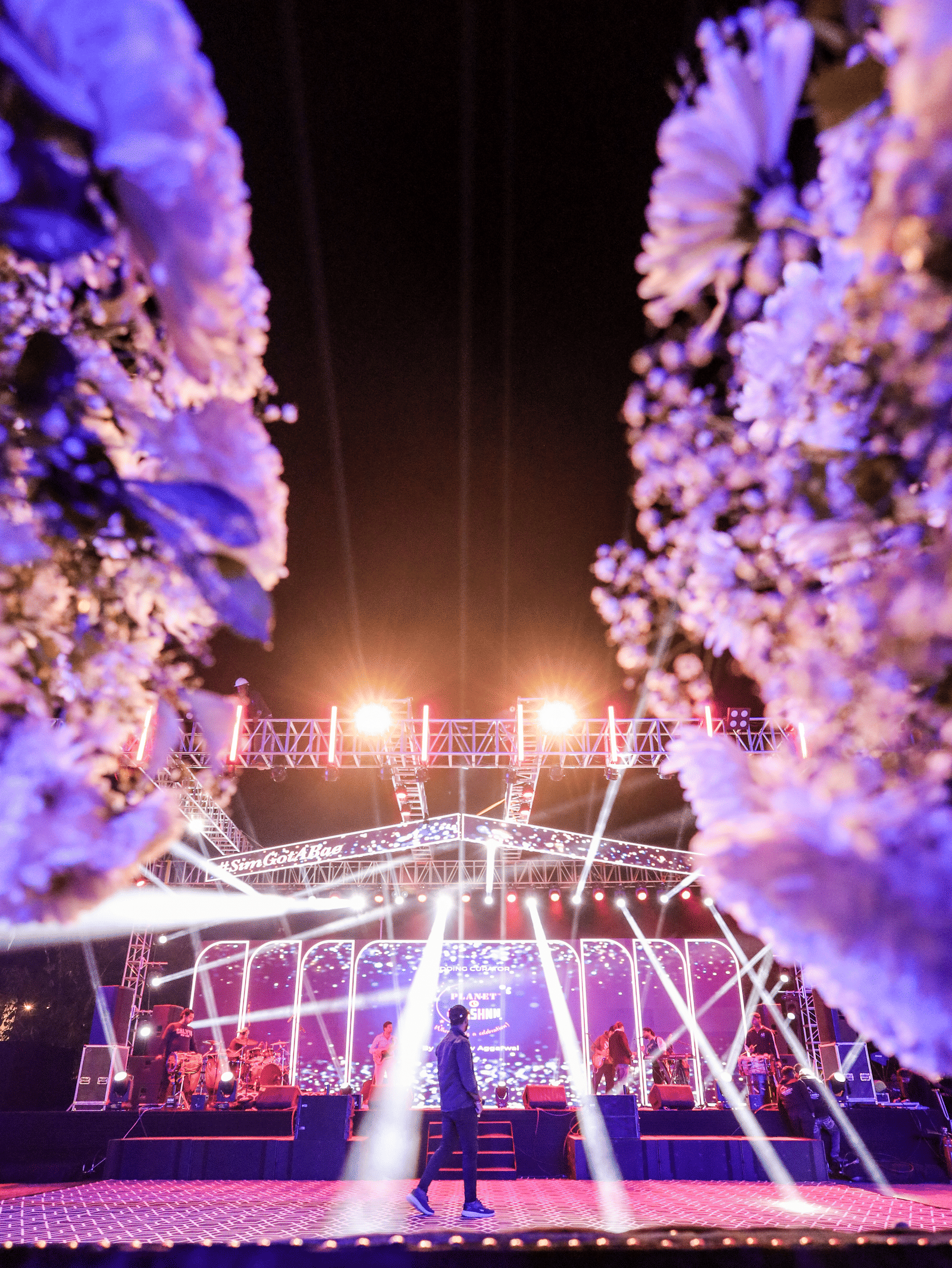 A view down a floral corridor toward a brightly lit performance stage at Ananta Spa & Resort, Jaipur, with one person standing.